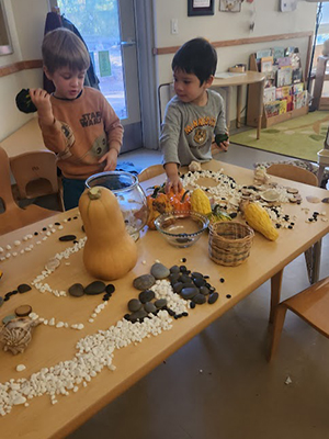 Two young children arrange many stones, gourds, squash, baskets and shells on a long table