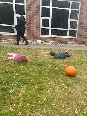 A large pumpkin rolls down a grassy hill followed by two children