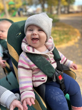 A smiling infant buckled into a cart for a walk outside, wearing a knitted cap and sweater