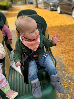 A smiling infant buckled into a cart for a walk outside, waving and wearing a green sweater and orange bandana.